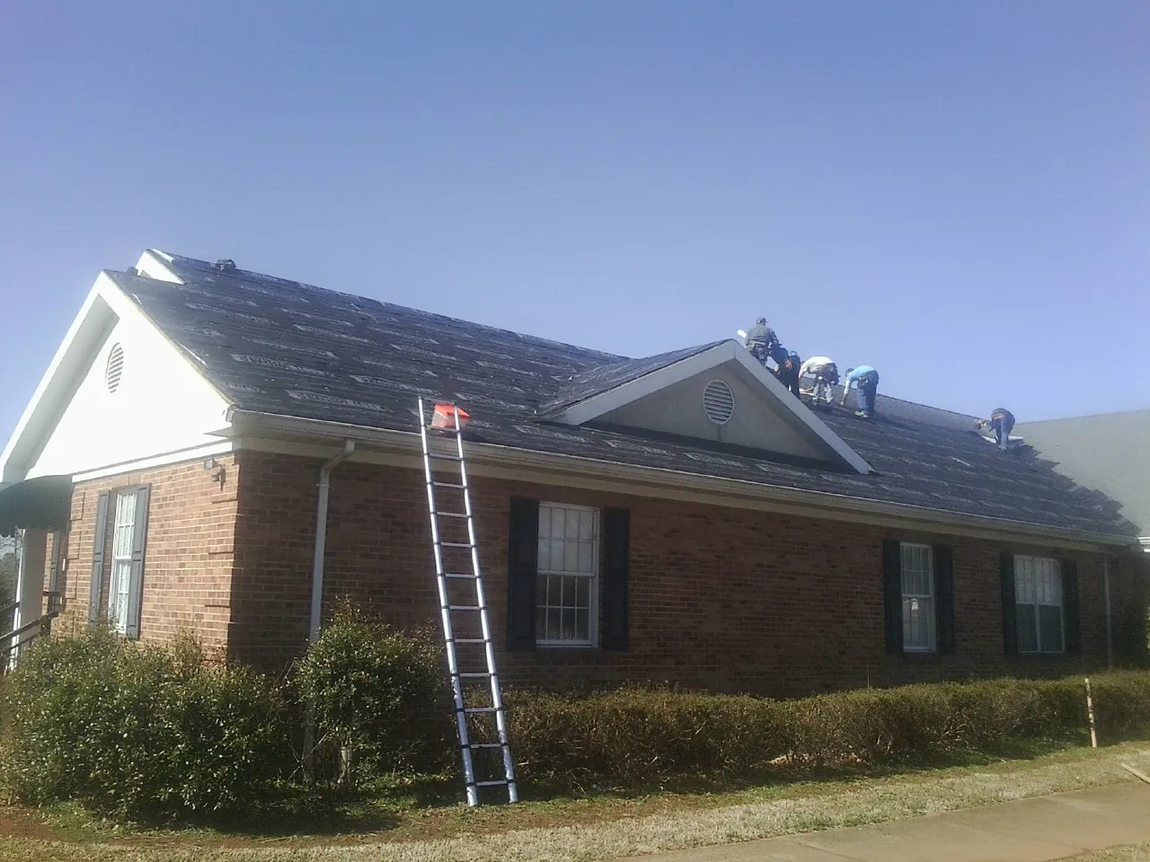 Crew tearing shingles off a brick ranch roof with a ladder set — Emergency Roof Repair work by our crew in Lavinia
