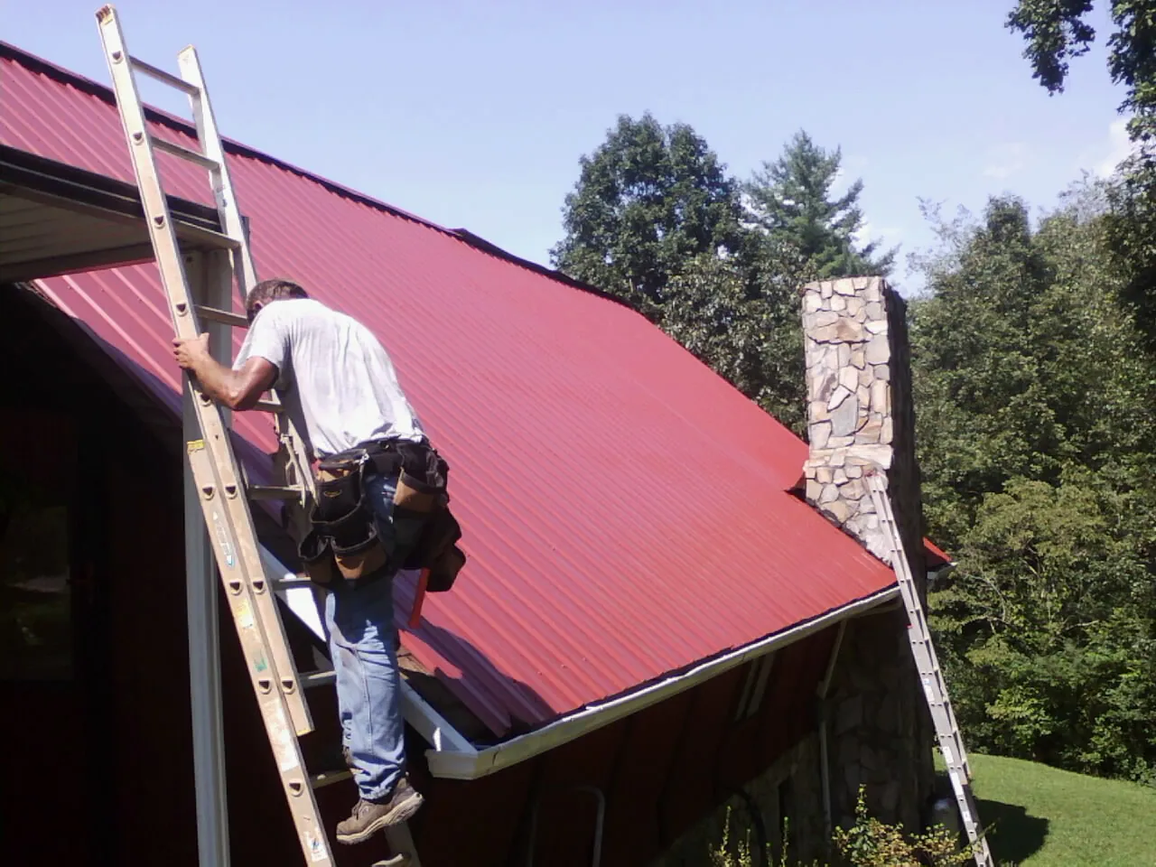 Roofer on a ladder beside a red metal roof and stone chimney — Soffit & Fascia Repair by Roofing Brothers in Lavinia
